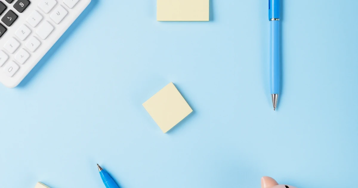 Abstract arrangement of calculator, sticky note pad, pen, and piggy bank on blue background.